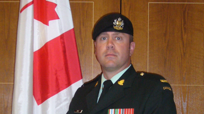 A man in the Canadian Armed Forces posed in front of a wood paneled wall, and a Canadian flag to his left. 