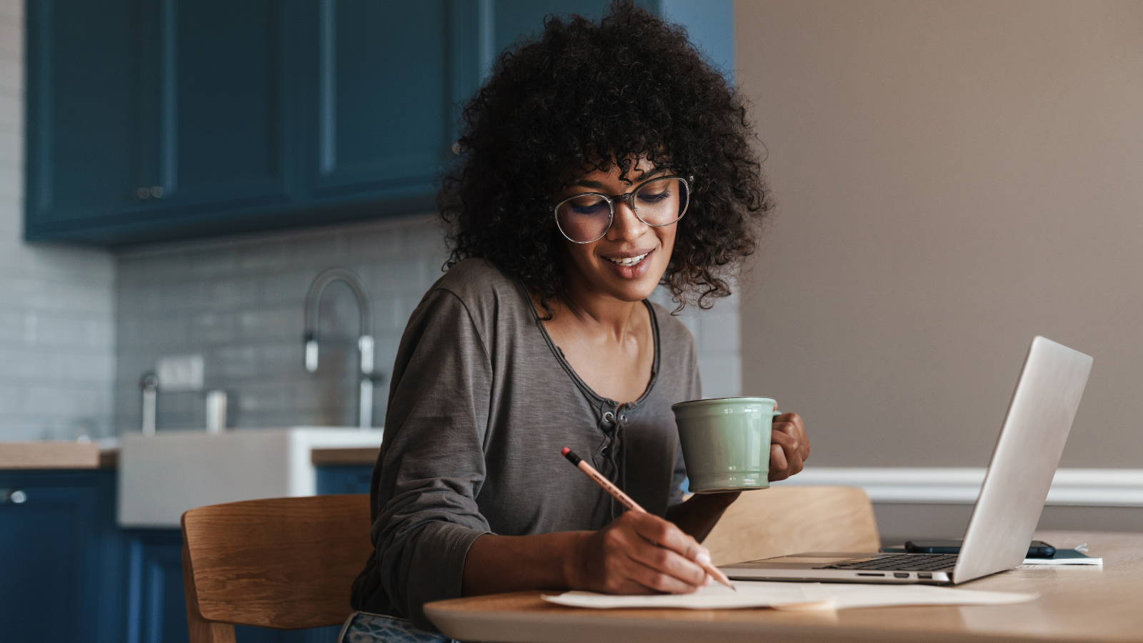 A woman using a laptop, holding a mug and taking notes. 