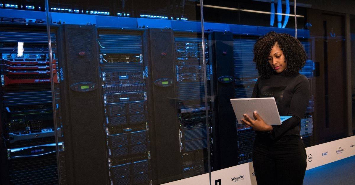A woman is standing in front of computers, while using a laptop.